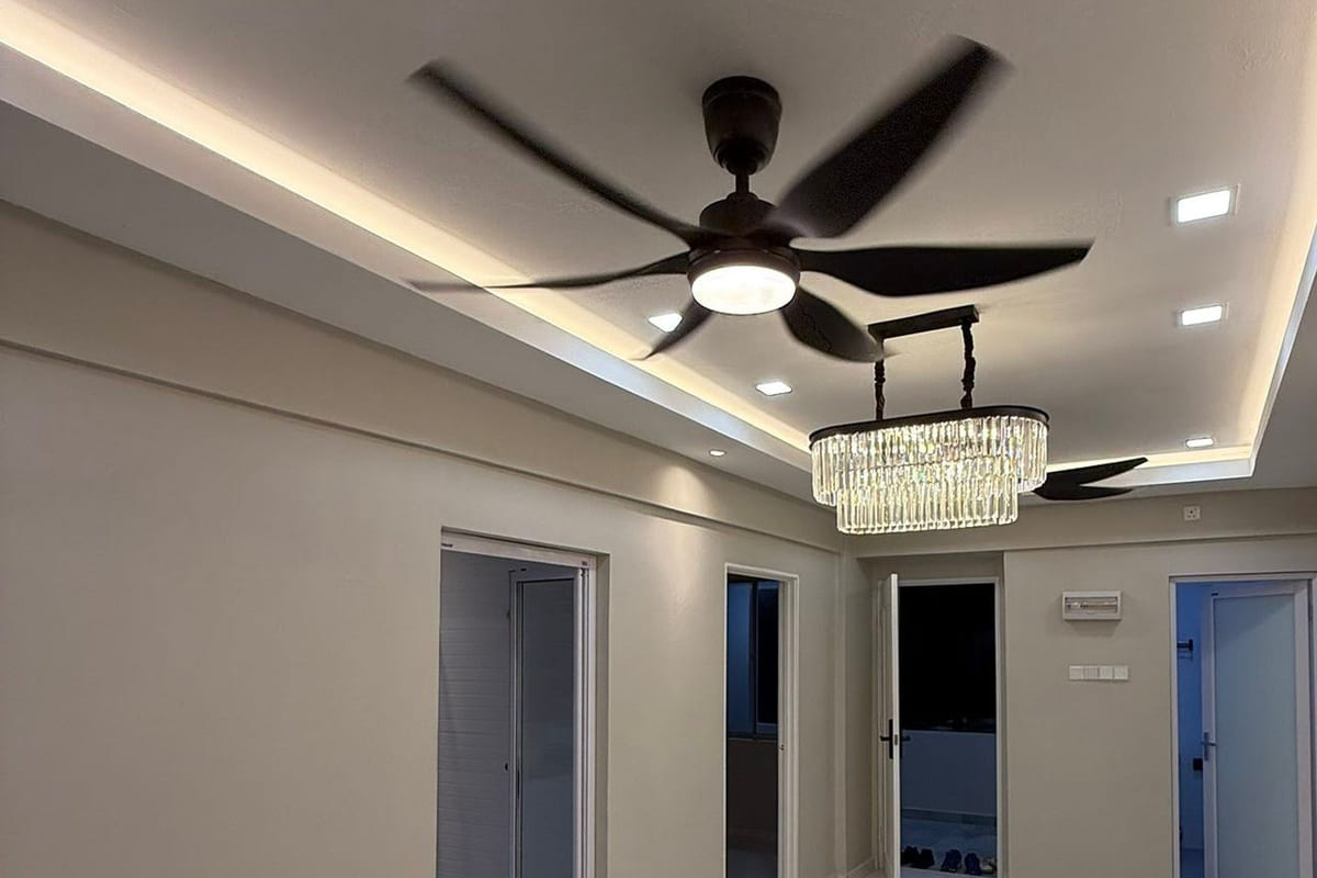 A living area looking down the length of a flat, under a stepped plaster cornice ceiling with a warm cove-lit reveal, a black ceiling fan and a rectangular crystal pendant.