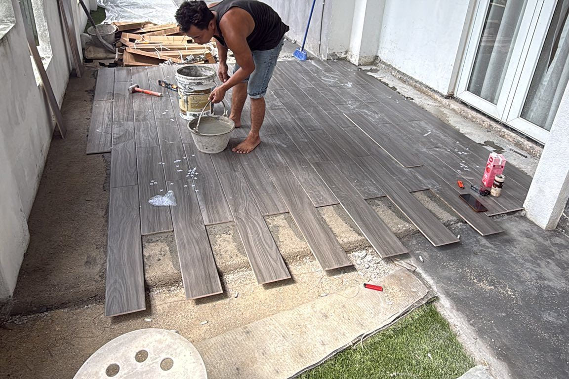 A tiler laying decking-style plank tiles on a new patio screed under a black steel patio frame with a translucent roof, beside a white house wall with sliding glazed doors.