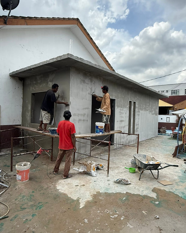 Three workers rendering new blockwork walls from a timber-plank staging on red steel trestles, in front of a flat-roofed extension.