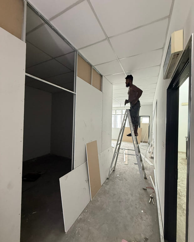 A worker on a step-ladder fixing a suspended ceiling, with plasterboard sheets leaning against the new partitions.