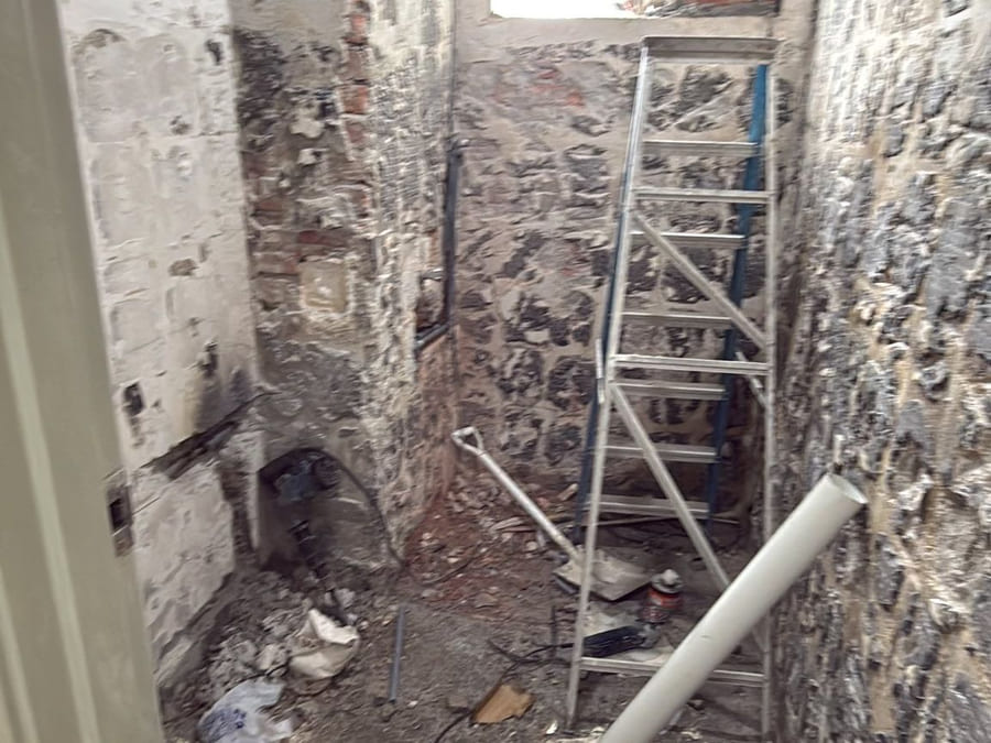 A stripped-out bathroom taken back to bare brick and screed, with a ladder, rubble and a length of new PVC waste pipe on the floor.